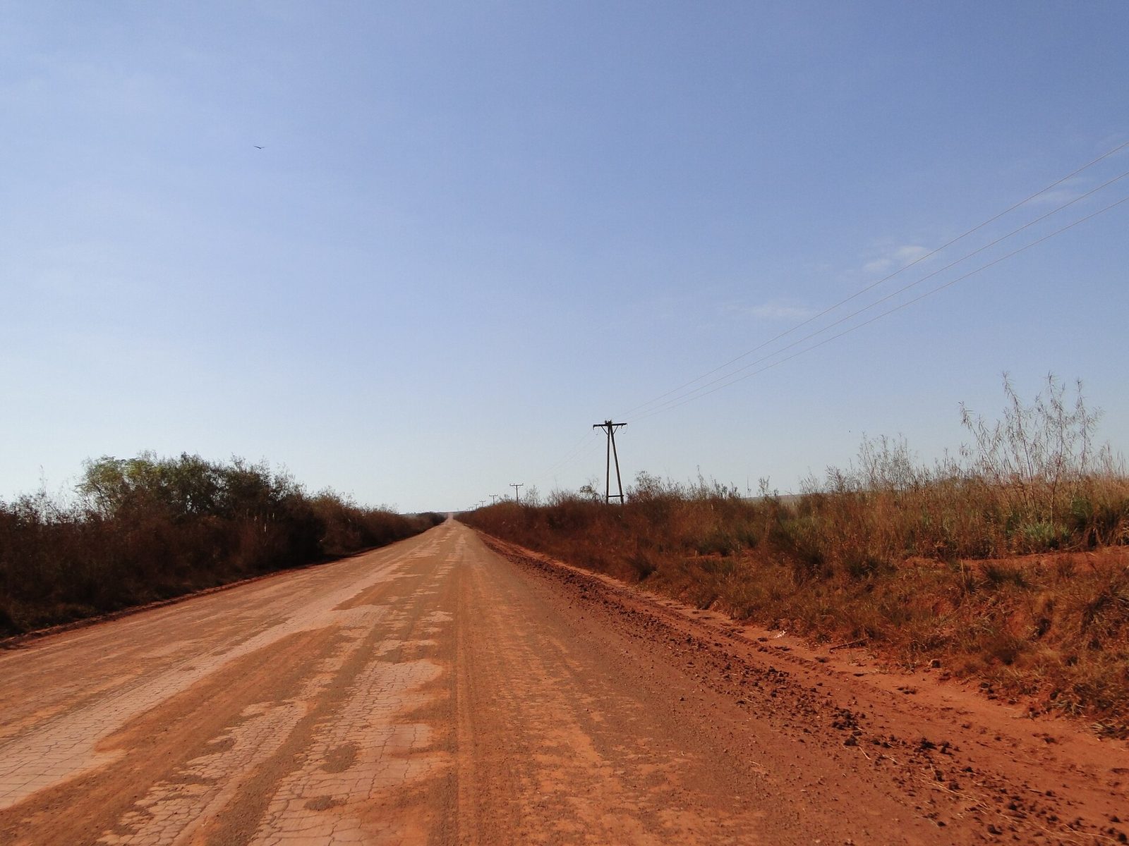 Straight red-earth road running between dry grass and scrub under a clear sky, with a power line beside it
