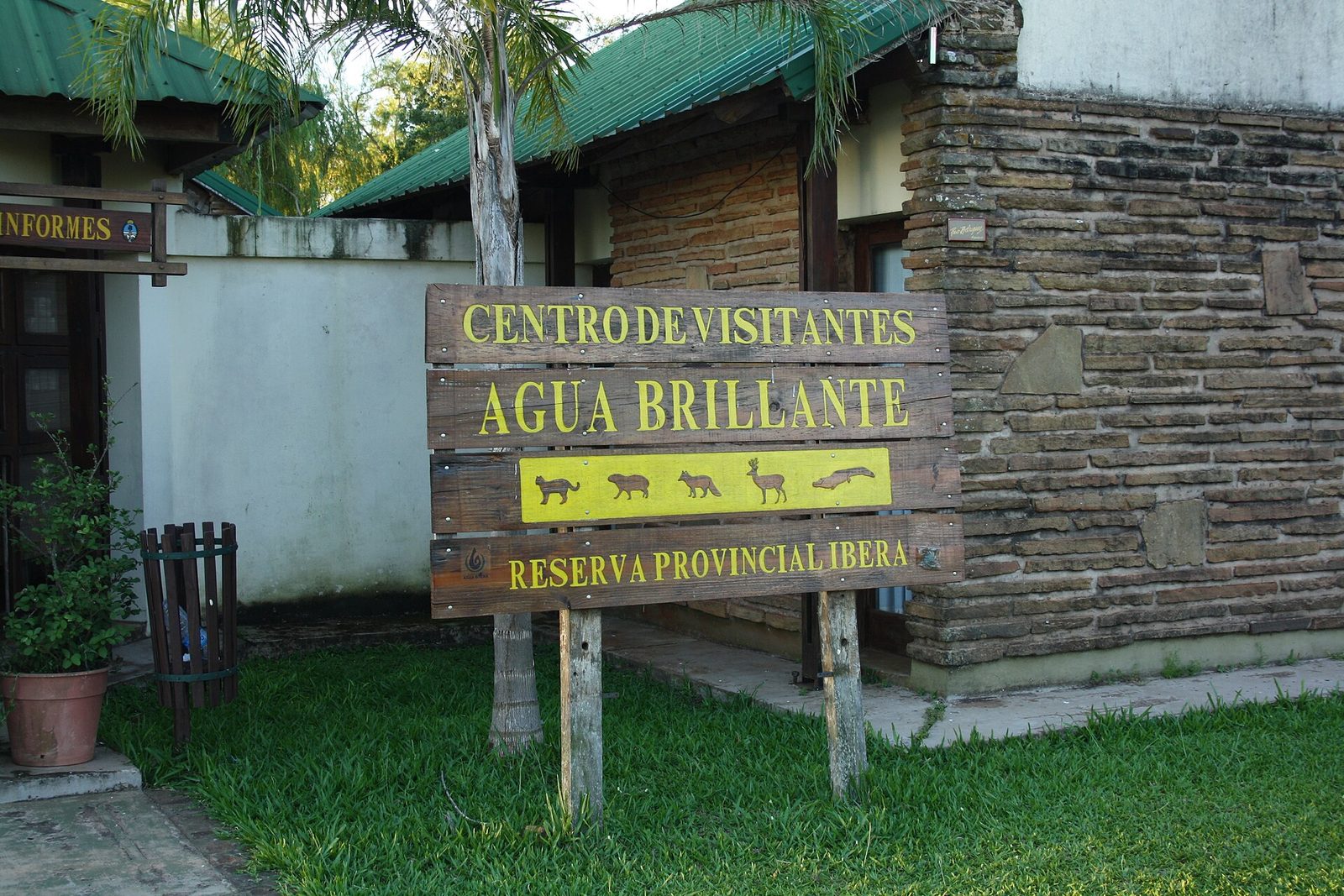 Wooden sign reading Centro de Visitantes Agua Brillante, Reserva Provincial Iberá, with animal silhouettes, in front of a stone building