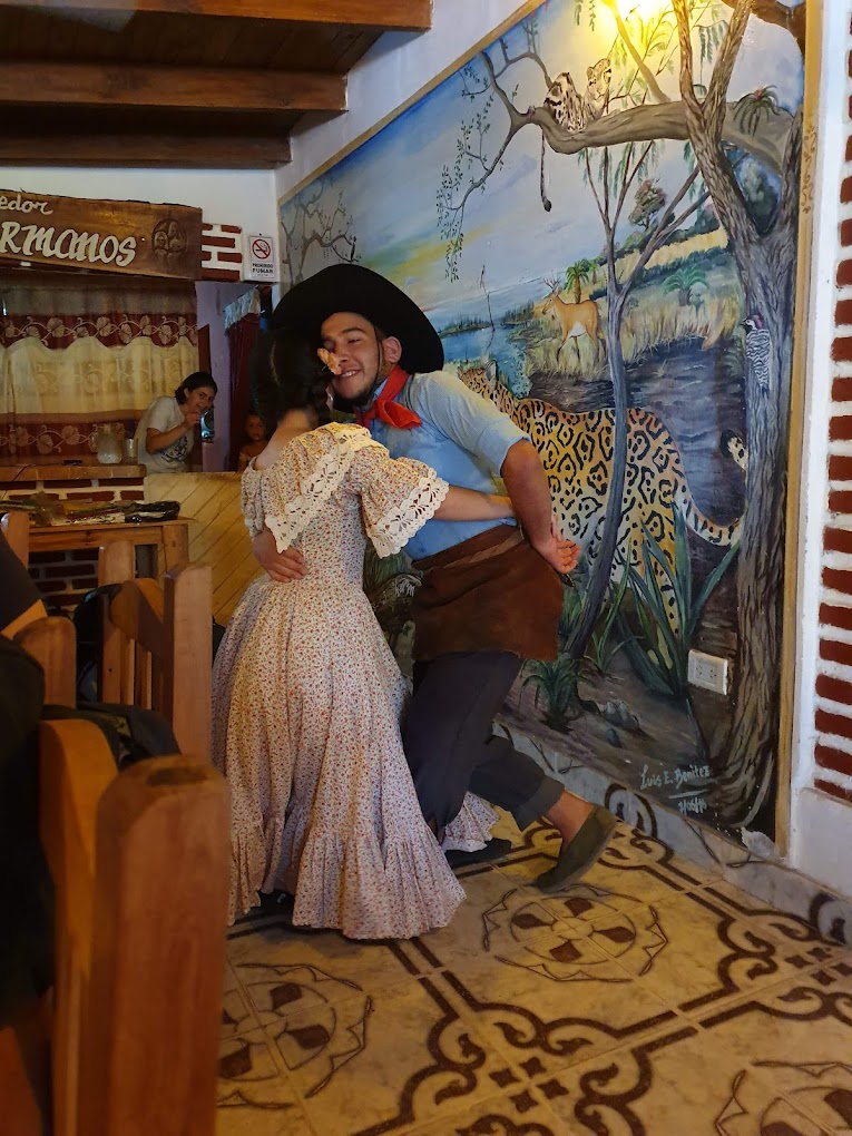 Pareja bailando folklore gaucho frente al mural del Iberá en Los 3 Hermanos