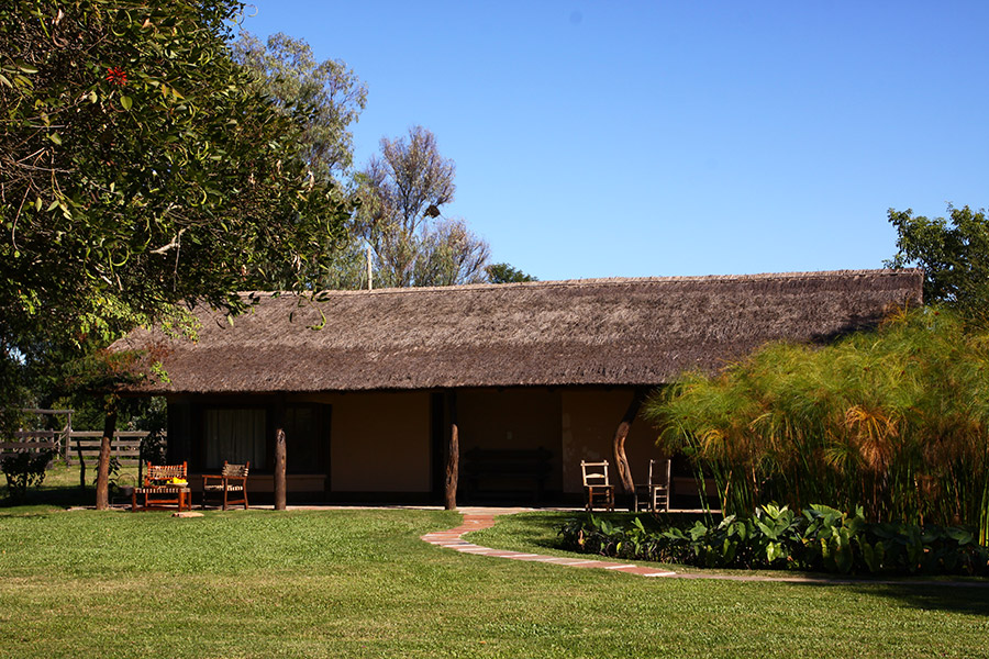 Fachada del rancho con techo de paja y galería  -  jardín bajo cielo azul